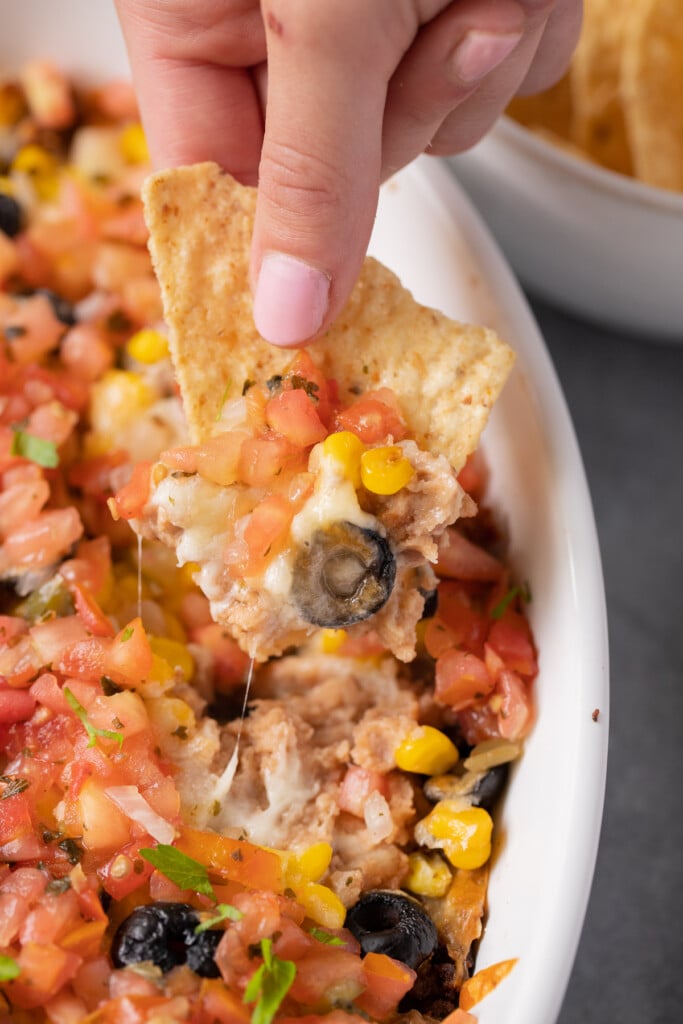 A hand and chip taking a scoop of dip out of baked seven layer dip from a white baking dish