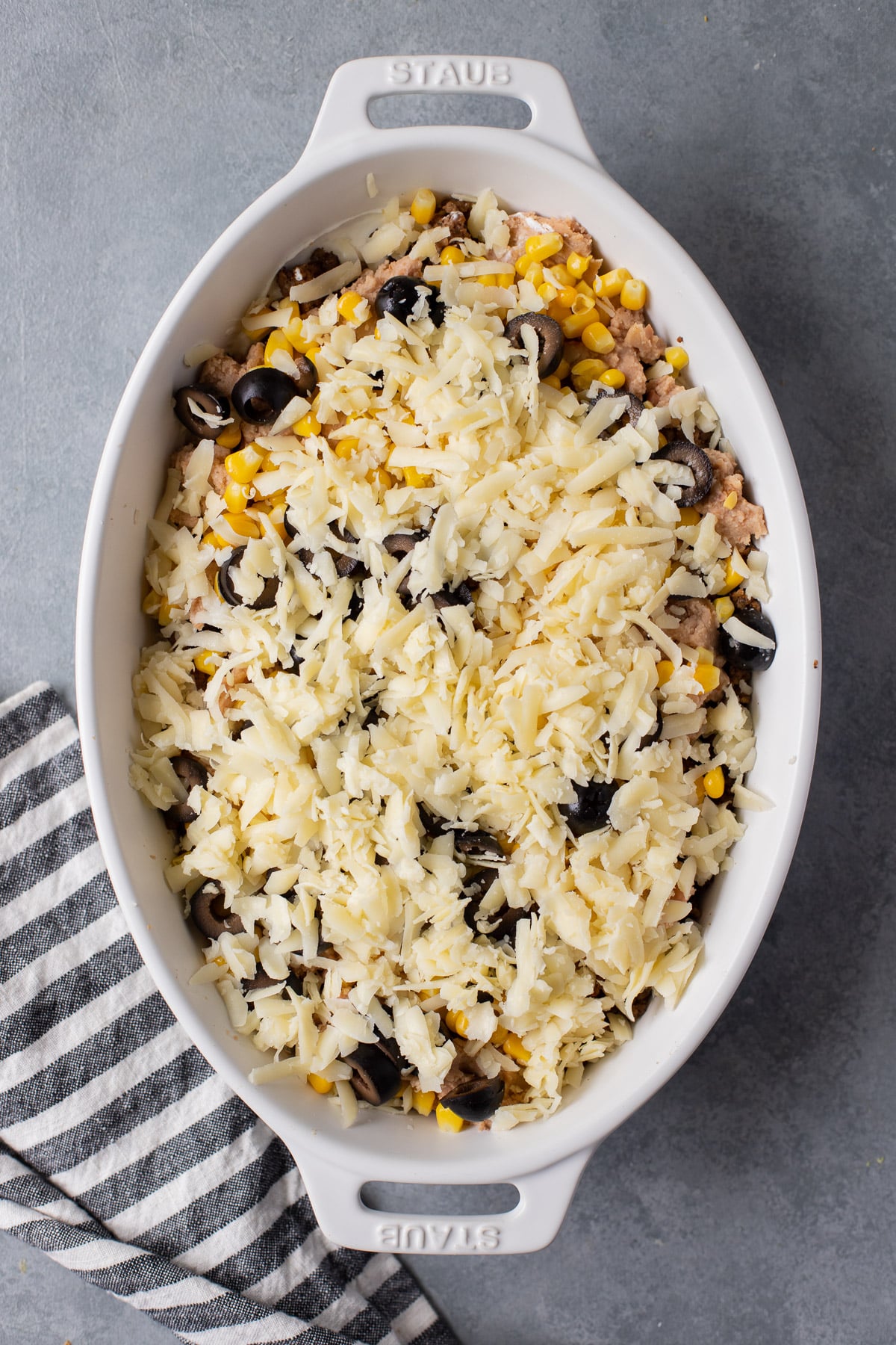 Layers of cream cheese, beans, corn, olives, cheese in a white baking dish for seven layer dip before baking