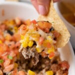 A hand and chip taking a scoop out of baked seven layer dip from a white baking dish