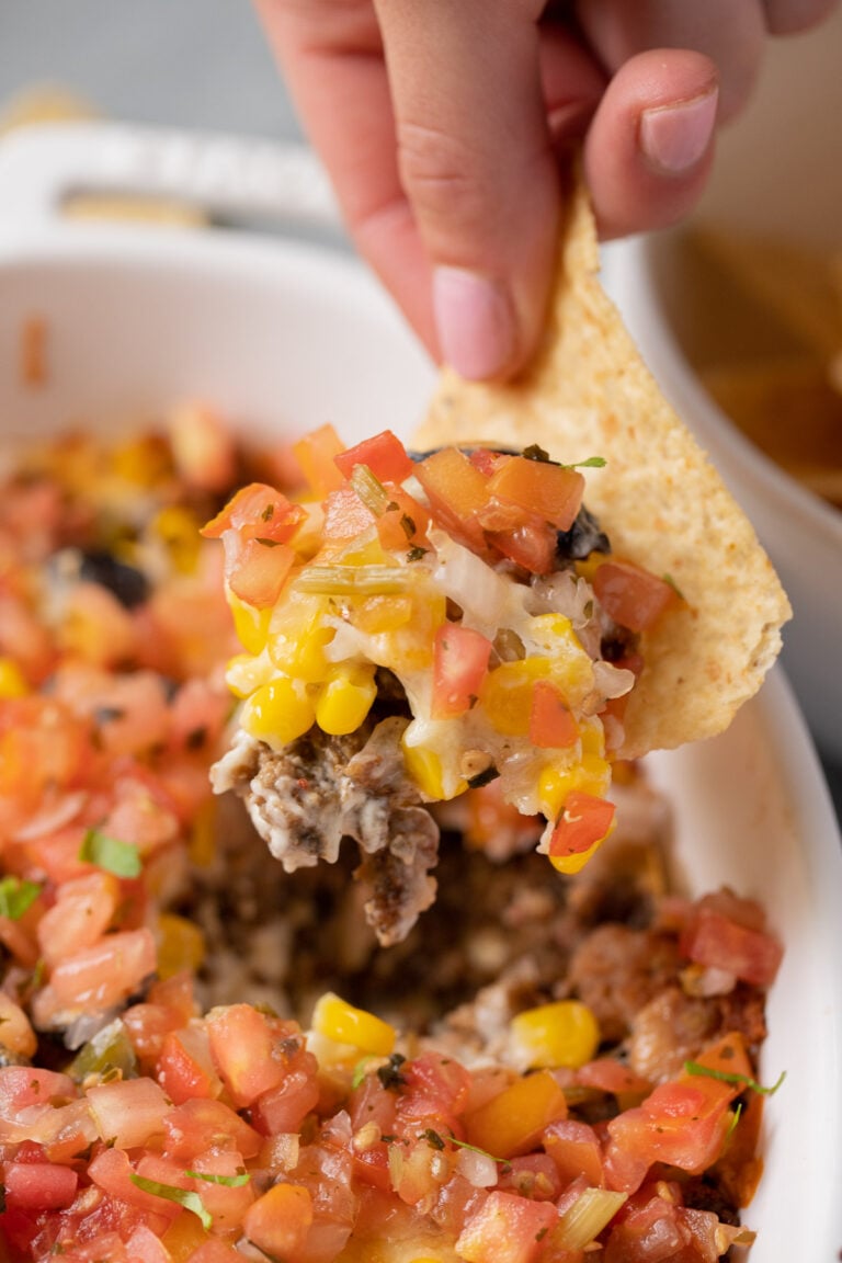 A hand and chip taking a scoop out of baked seven layer dip from a white baking dish