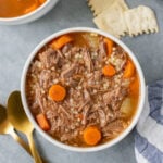 A white bowl of pot roast soup with carrot coins on a grey background with gold spoons, crackers and another bowl on the side