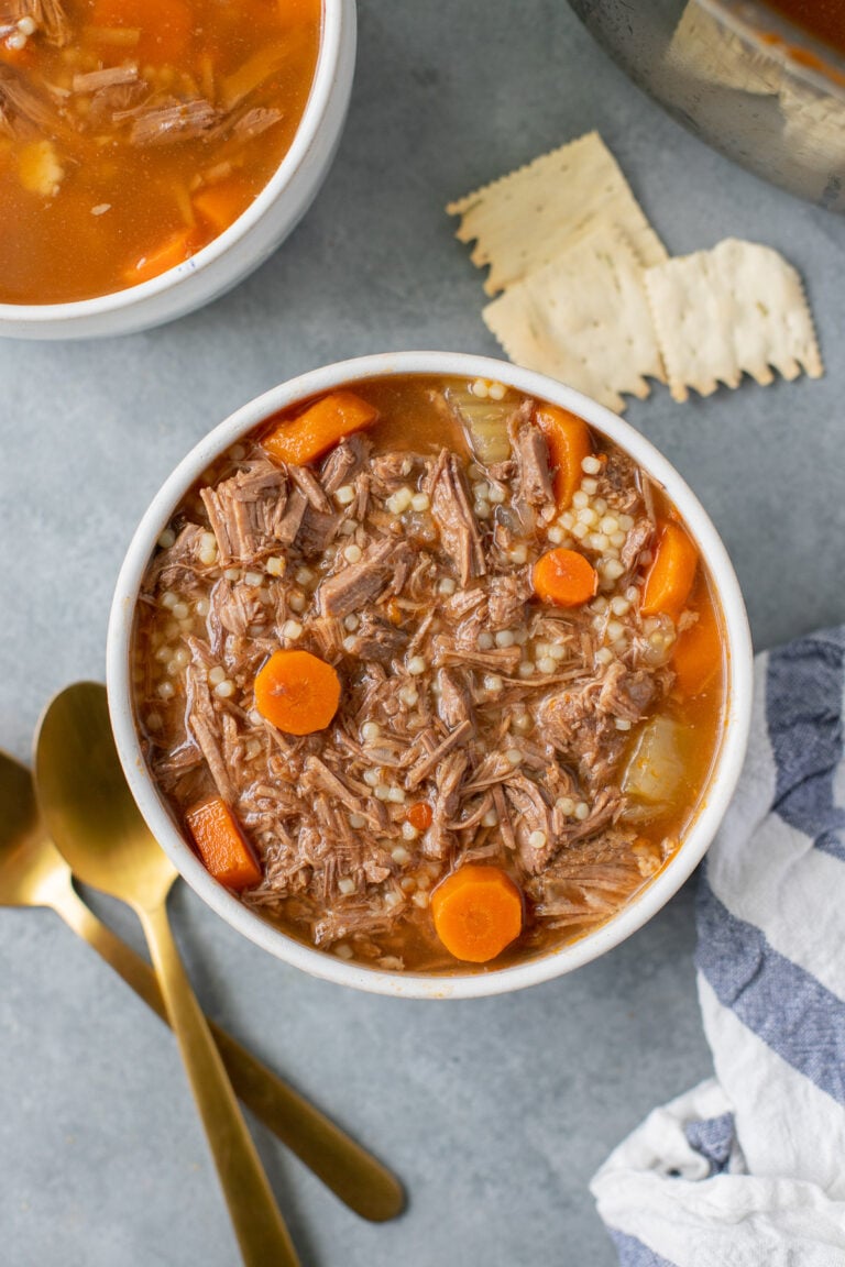 A white bowl of pot roast soup with carrot coins on a grey background with gold spoons, crackers and another bowl on the side