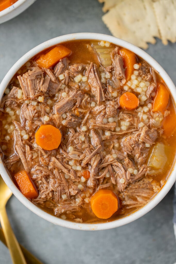A close up of a white bowl of pot roast soup with carrot coins on a grey background
