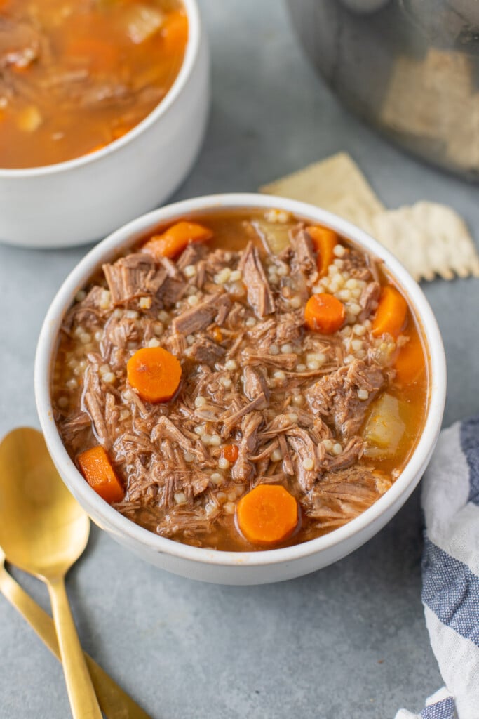 A white bowl of pot roast soup with carrot coins on a grey background with gold spoons, crackers and another bowl of soup on the side