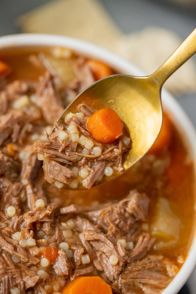 A gold spoon taking a spoonful out of a white bowl of pot roast soup with crackers in the background