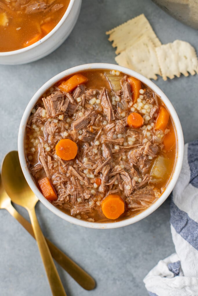 A bowl of pot roast soup with carrot coins on a grey background with gold spoons, crackers and another bowl on the side