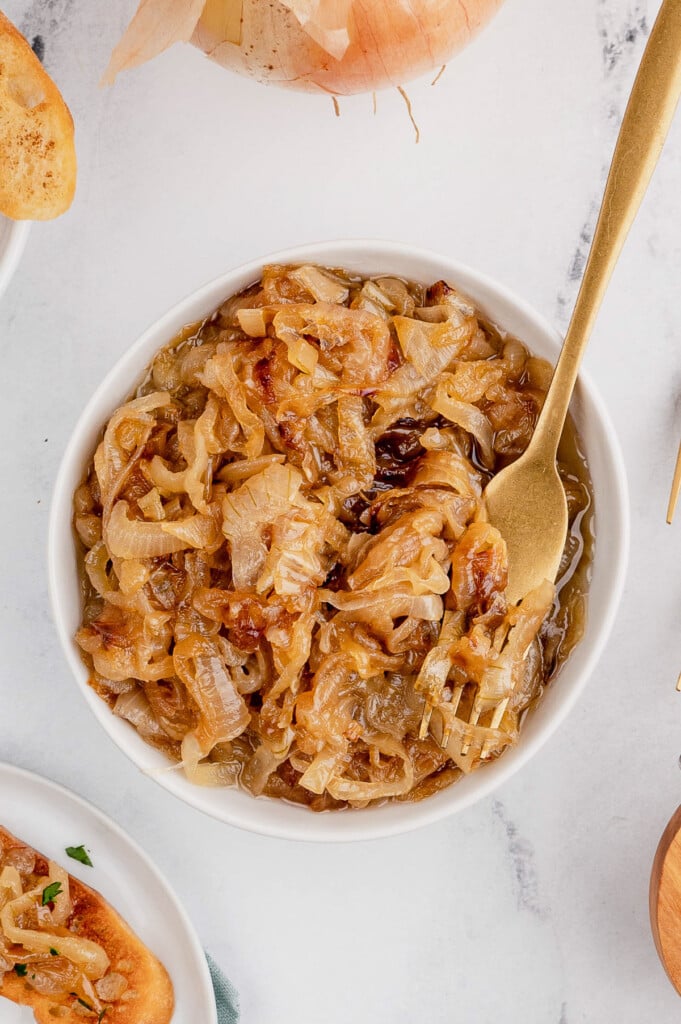 An overhead shot of a white bowl filled with caramelized onions with a gold fork in it on a marble background
