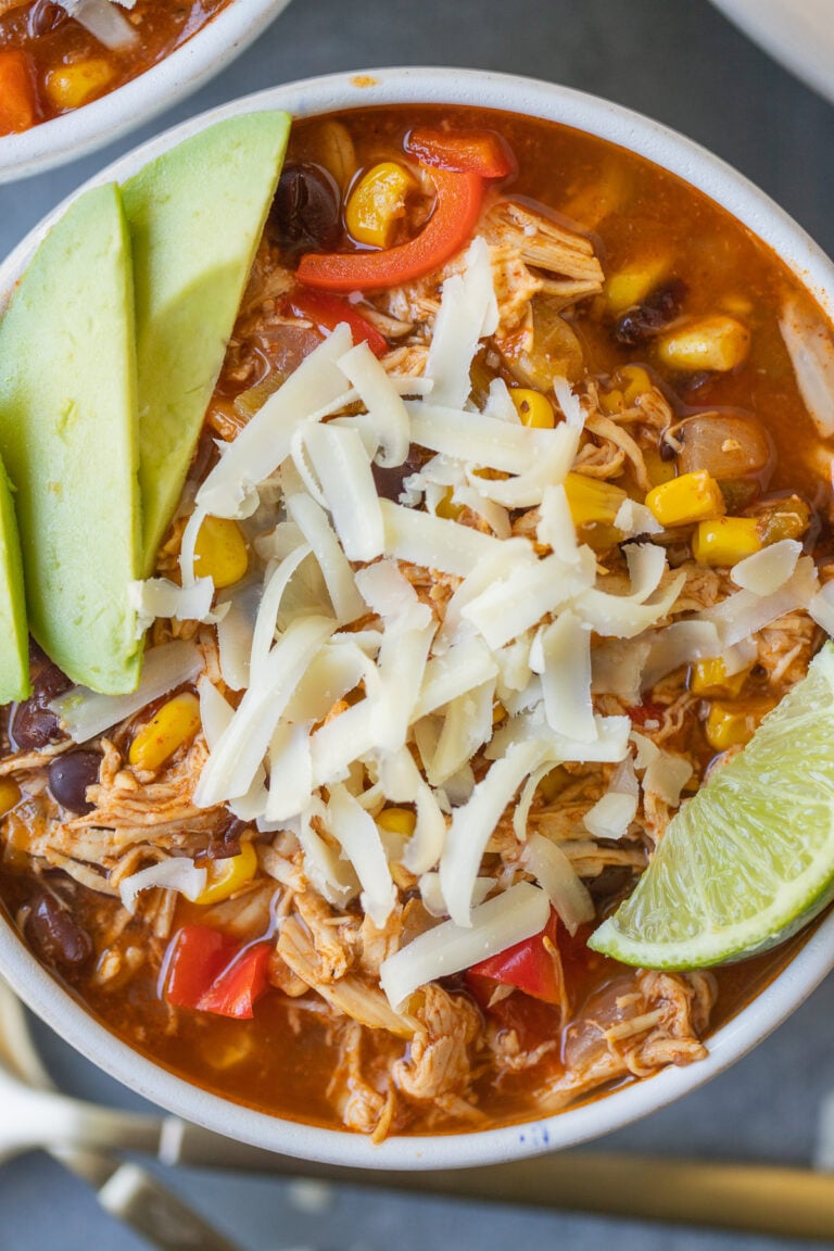 A close up of Chicken Enchilada Soup in a white bowl with avocado, lime and cheese on top