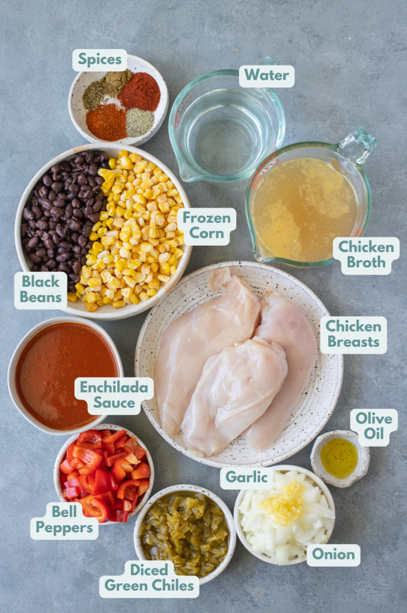 Ingredients for Enchilada Soup in small white bowls on a grey background: olive oil, onion, garlic, bell peppers, boneless skinless chicken breast, homemade enchilada sauce, diced green chiles, chicken broth, paprika, pepper, ground cumin, chili powder, oregano, black beans, frozen corn
