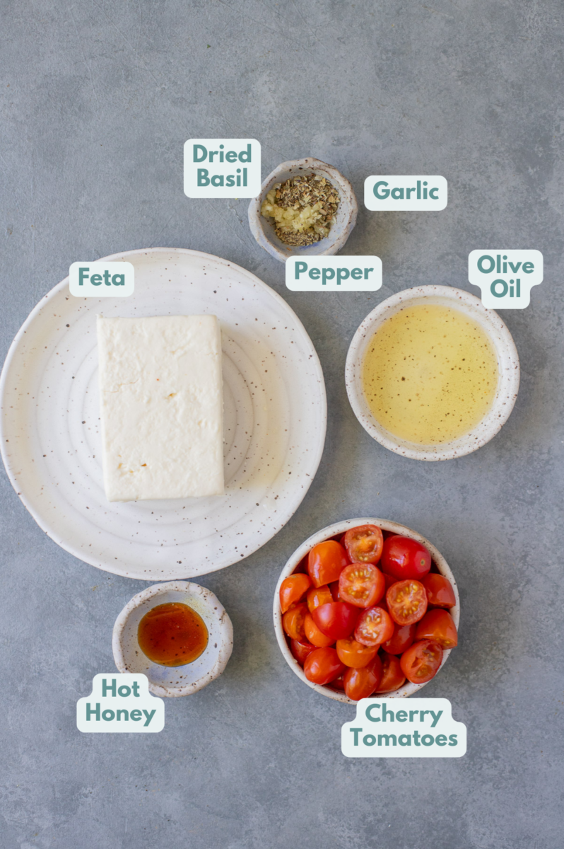 Ingredients for baked feta dip in white bowls: feta, olive oil, cherry tomatoes, garlic, dried basil, pepper, hot honey