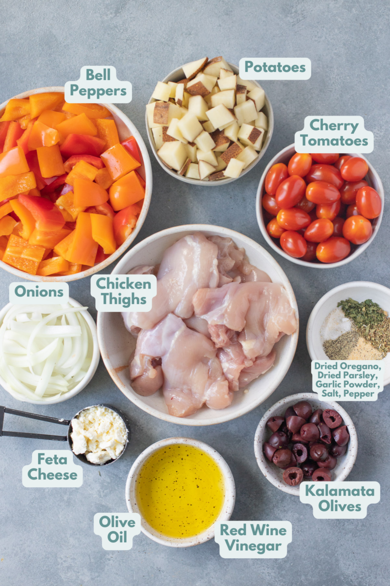 Ingredients for greek chicken thighs in white bowls: chicken thighs, bell peppers, potatoes, cherry tomatoes, kalamata olives, onions, crumbled feta (optional), olive oil, red wine vinegar, garlic powder, dried oregano, dried parsley, salt & pepper