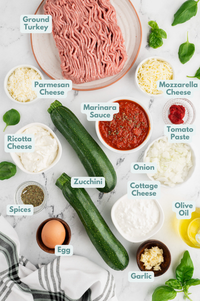 Ingredients for easy zucchini lasagna in white bowls: zucchini, ground turkey, onion, garlic, tomato paste, marinara sauce, mozzarella cheese, parmesan cheese, olive oil, dried basil, dried parsley, dried oregano, fresh basil, salt & pepper, ricotta cheese, cottage cheese, egg, dried basil, dried parsley, dried oregano