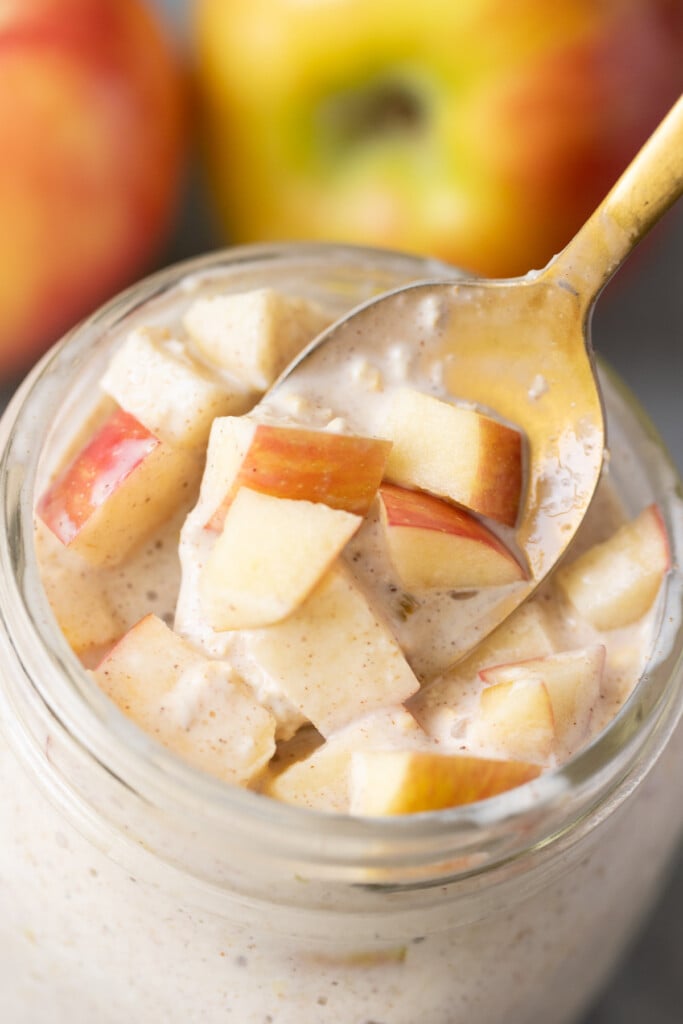 A gold spoon taking a bite out of a jar of apple overnight oats with apples in the backgrouns