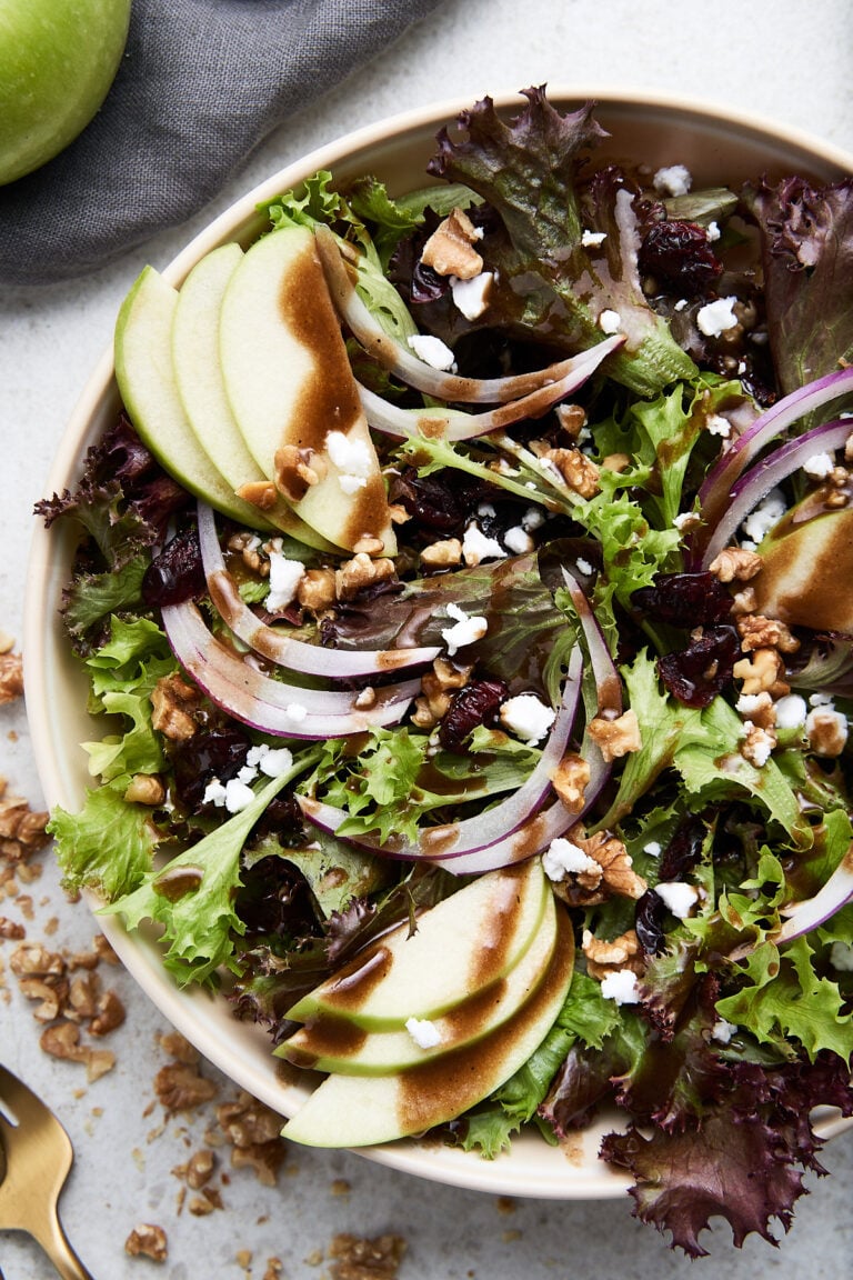 An overhead shot of an apple walnut salad with vinaigrette on top in a white bowl on a white background