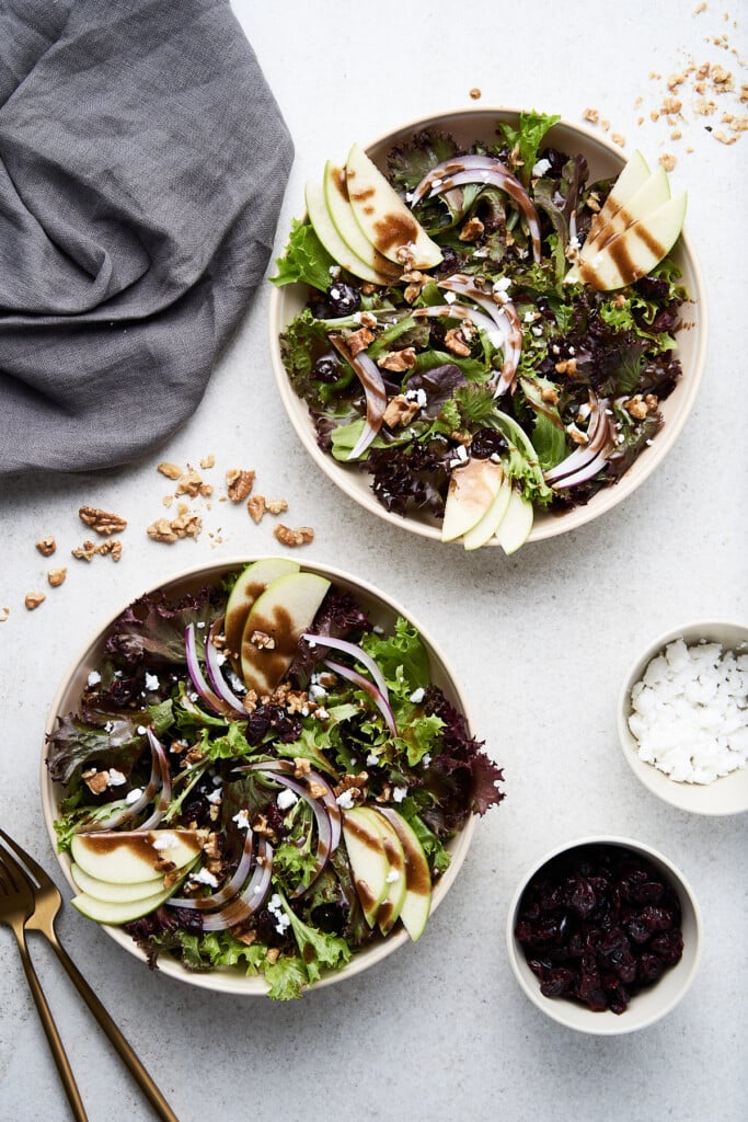 An overhead shot of two white bowls of apple walnut salad with two bowls of cranberries and feta cheese, a cloth and gold forks on the side