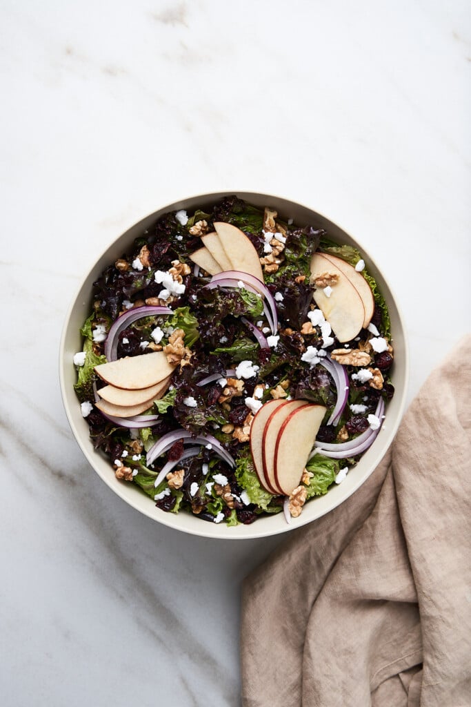 An overhead shot of a white bowl of apple walnut salad on a marble background and a tan cloth on the side