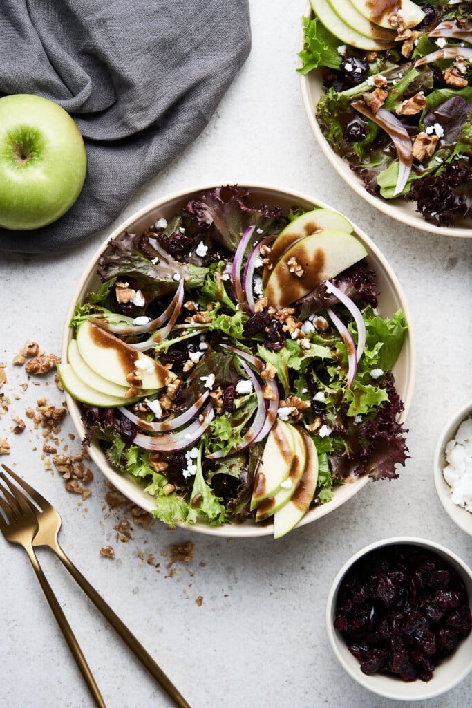 An overhead shot of a white bowl of apple walnut salad with cranberries, an apple, a cloth, and gold forks on the side