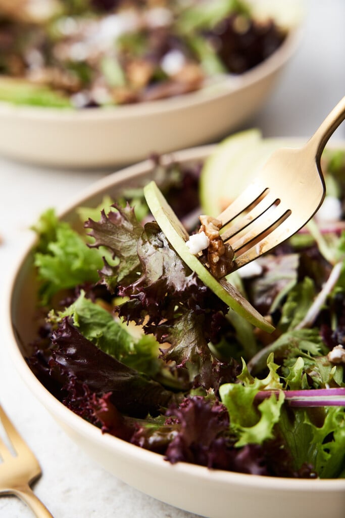 A gold fork taking a bite of apple walnut salad from a white bowl with another bowl and gold fork on the side