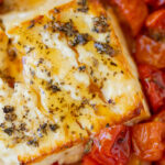 A block of baked feta cheese with herbs and hot honey on top with cherry tomatoes surrounding it