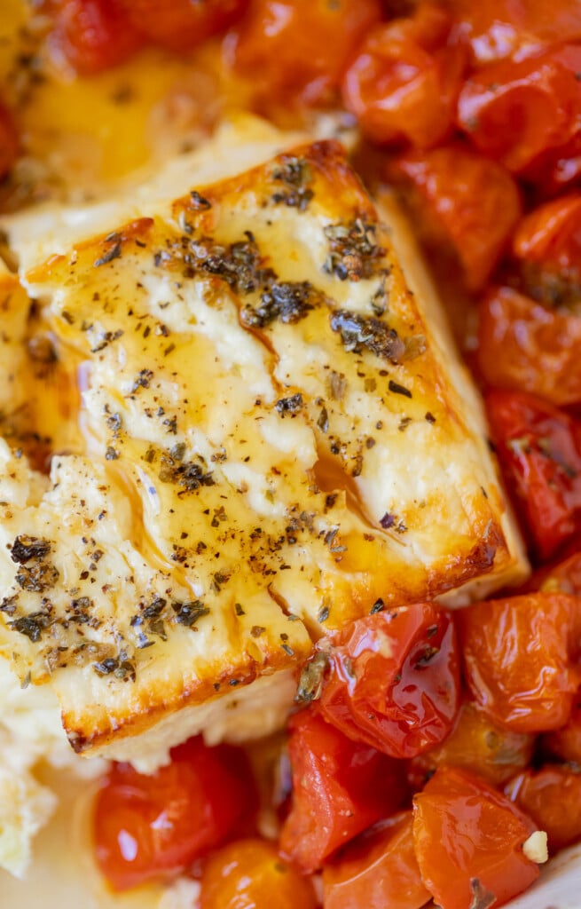 A block of baked feta cheese with herbs and hot honey on top with cherry tomatoes surrounding it