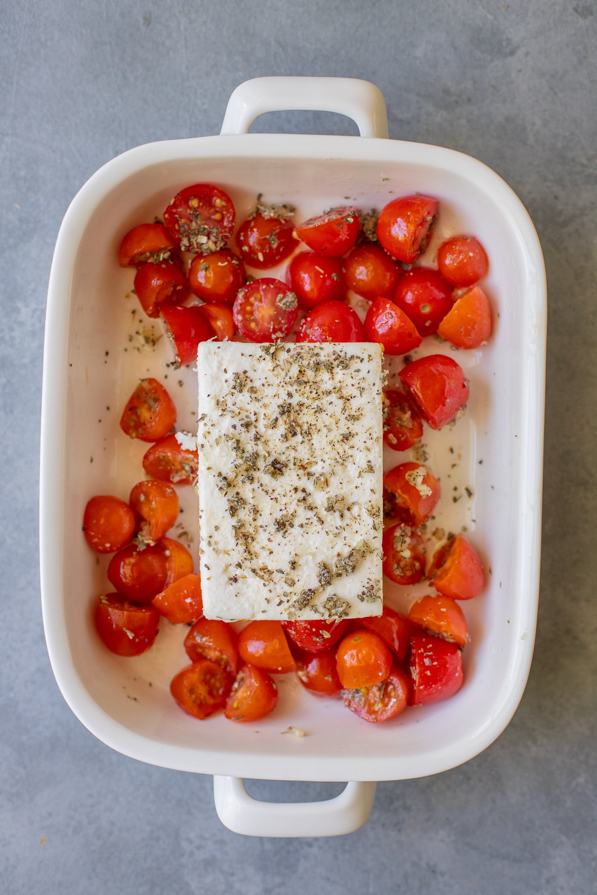An overhead shot of a white baking dish with a block of feta cheese in the middle surrounded by cherry tomatoes