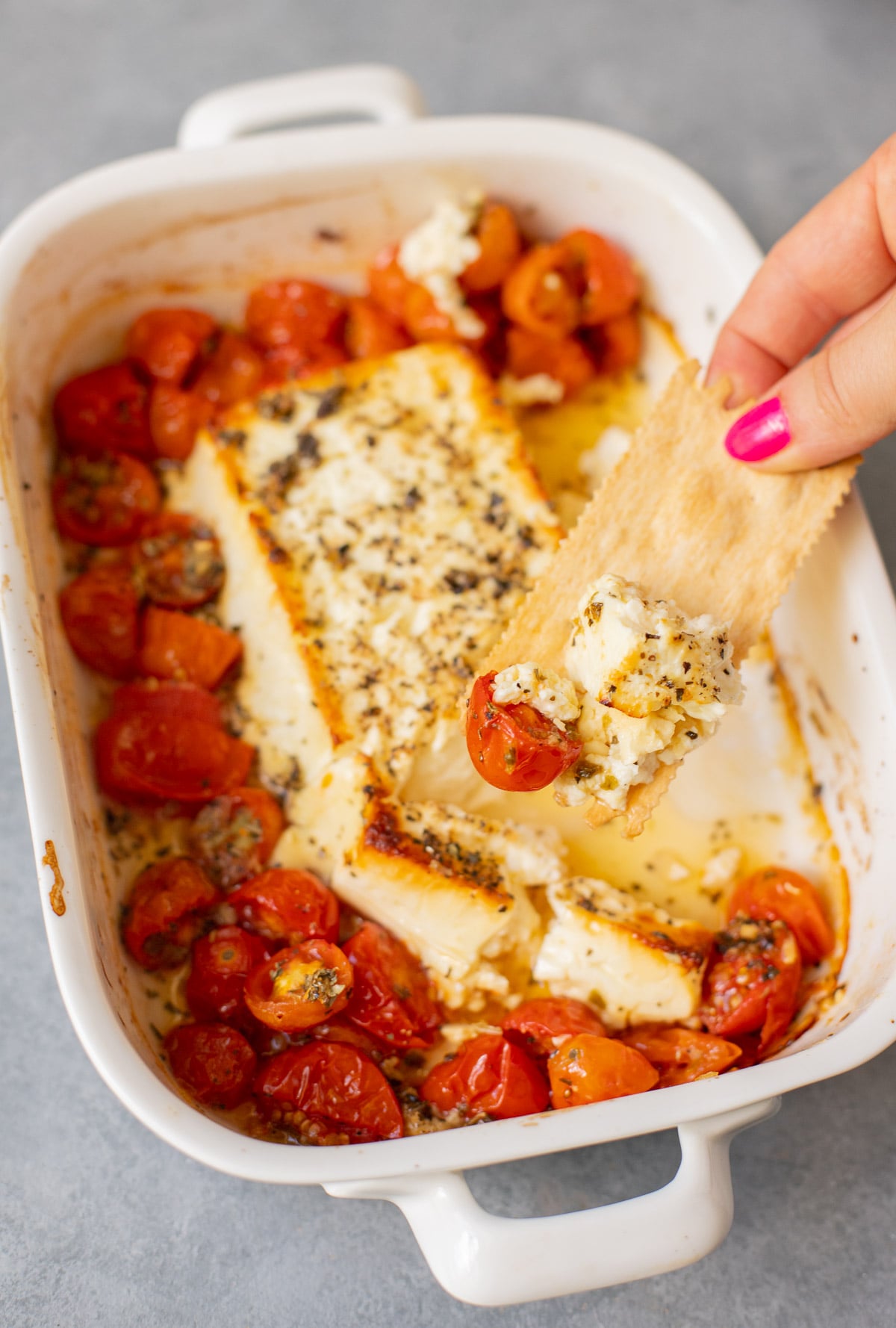 A white baking dish with baked feta dip with cherry tomatoes and a cracker taking a scoop from it