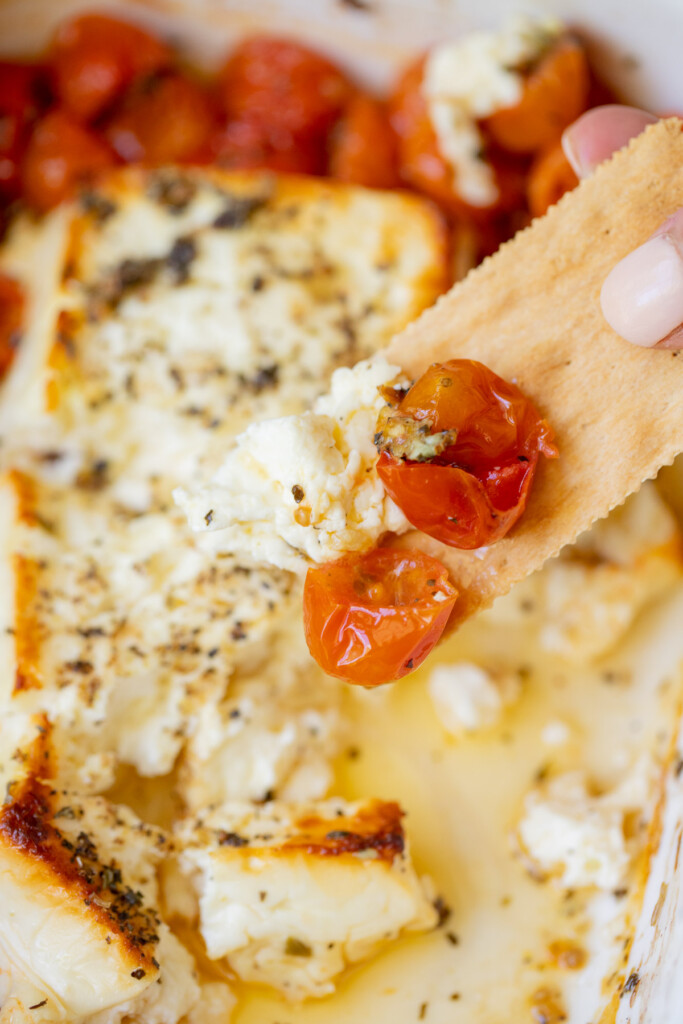 A cracker taking a scoop of baked feta dip with cherry tomatoes and the baking dish of the dip in the background