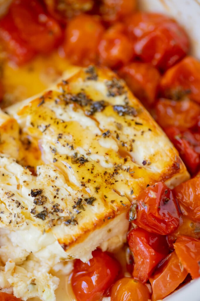 A block of baked feta cheese with herbs and hot honey on top with cherry tomatoes surrounding it