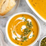An overhead shot of a white bowl of instant pot butternut squash soup with cream, herbs and pumpkin seeds on top with a dish of soup and bread on the side