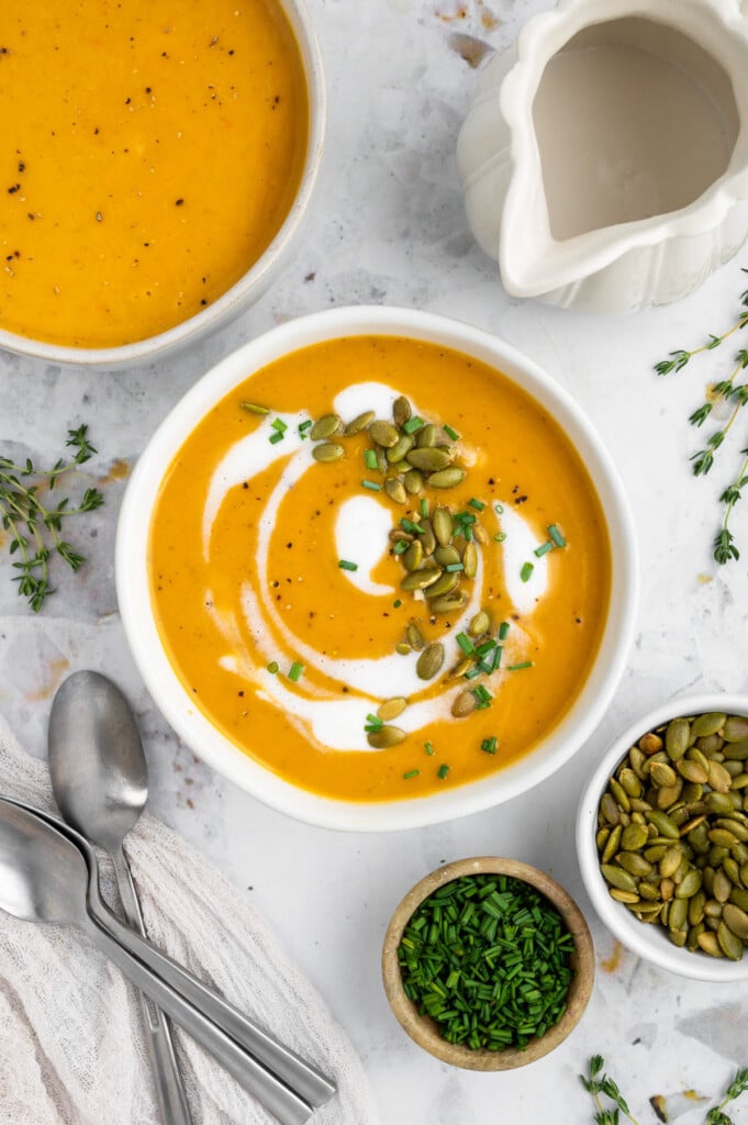 An overhead shot of a white bowl of instant pot butternut squash soup with cream, herbs and pumpkin seeds on top with a bowl of soup and herbs on the side