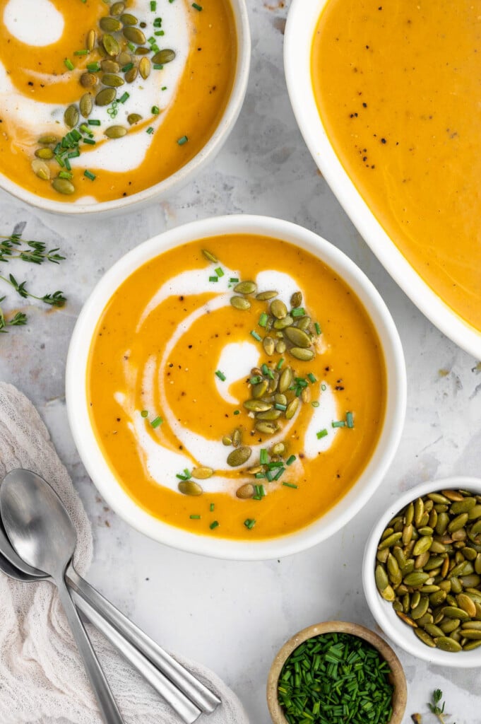 An overhead shot of a white bowl of instant pot butternut squash soup with cream, herbs and pumpkin seeds on top with a dish and bowl of soup on the side