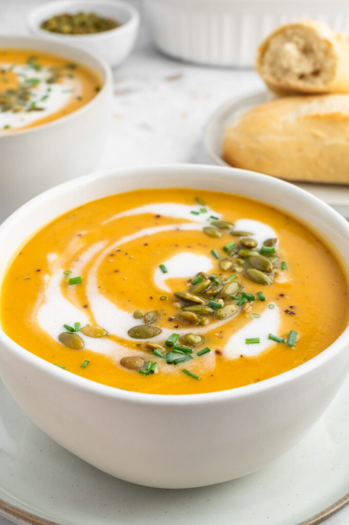 a bowl of instant pot butternut squash soup with cream and herbs on top with a bowl of soup and bread in the background