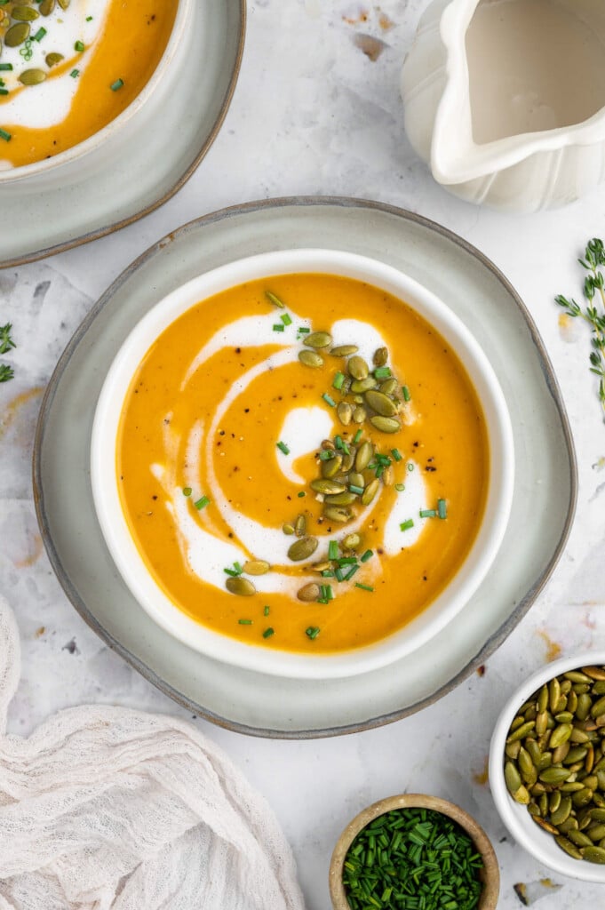 An overhead shot of a white bowl of instant pot butternut squash soup with cream, herbs and pumpkin seeds on top on a white background