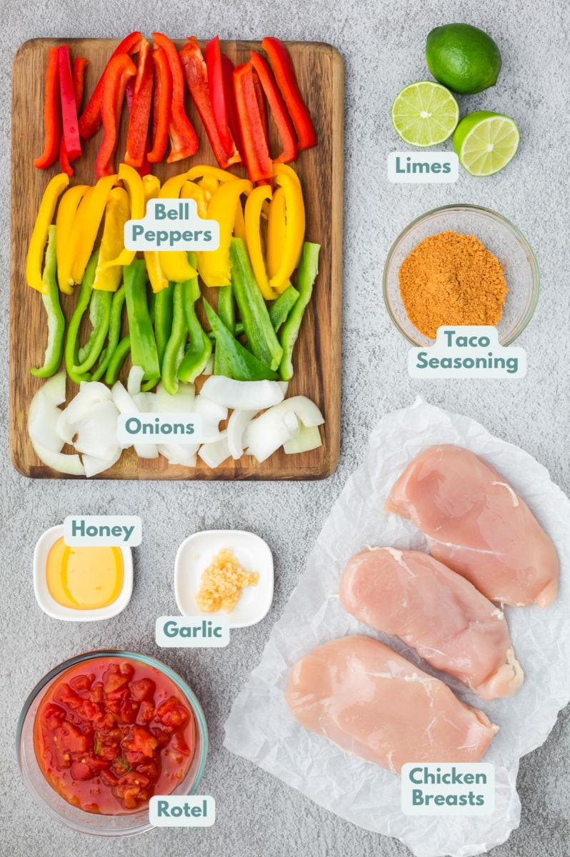 Ingredients for crock pot chicken fajitas laid on a grey background: chicken breasts, onion, red, yellow, and green pepper, Rotel, taco seasoning, honey, garlic, limes for fresh lime juice