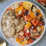 Greek Chicken Thighs and veggies on a white plate with gold forks and a sheet pan on the side
