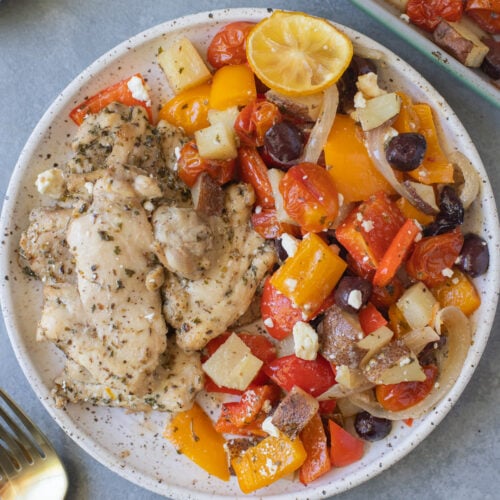 Greek Chicken Thighs and veggies on a white plate with gold forks and a sheet pan on the side
