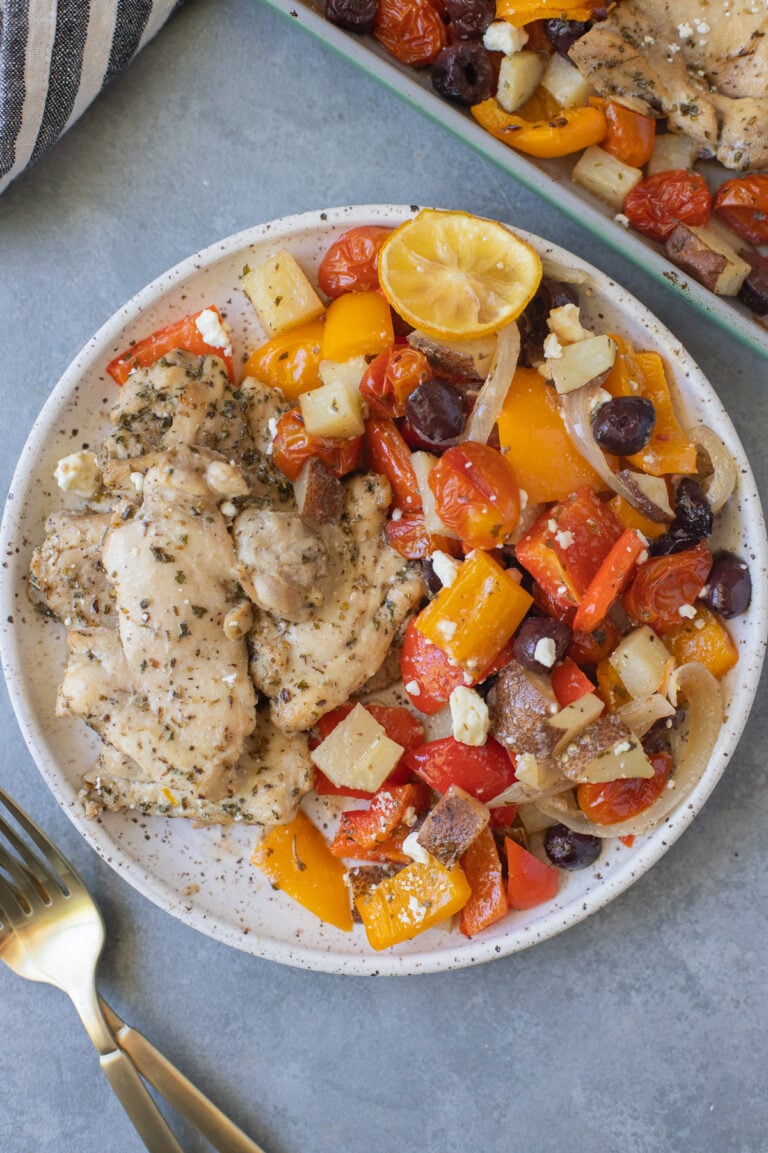 Greek Chicken Thighs and veggies on a white plate with gold forks and a sheet pan on the side