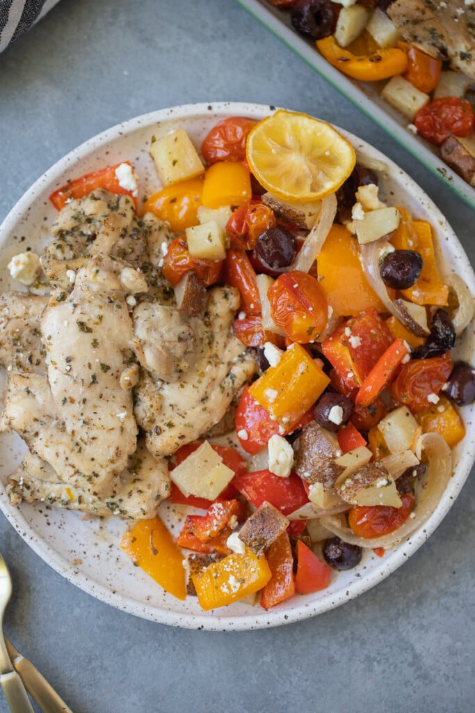A close up of Greek Chicken Thighs and veggies on a white plate with a sheet pan on the side
