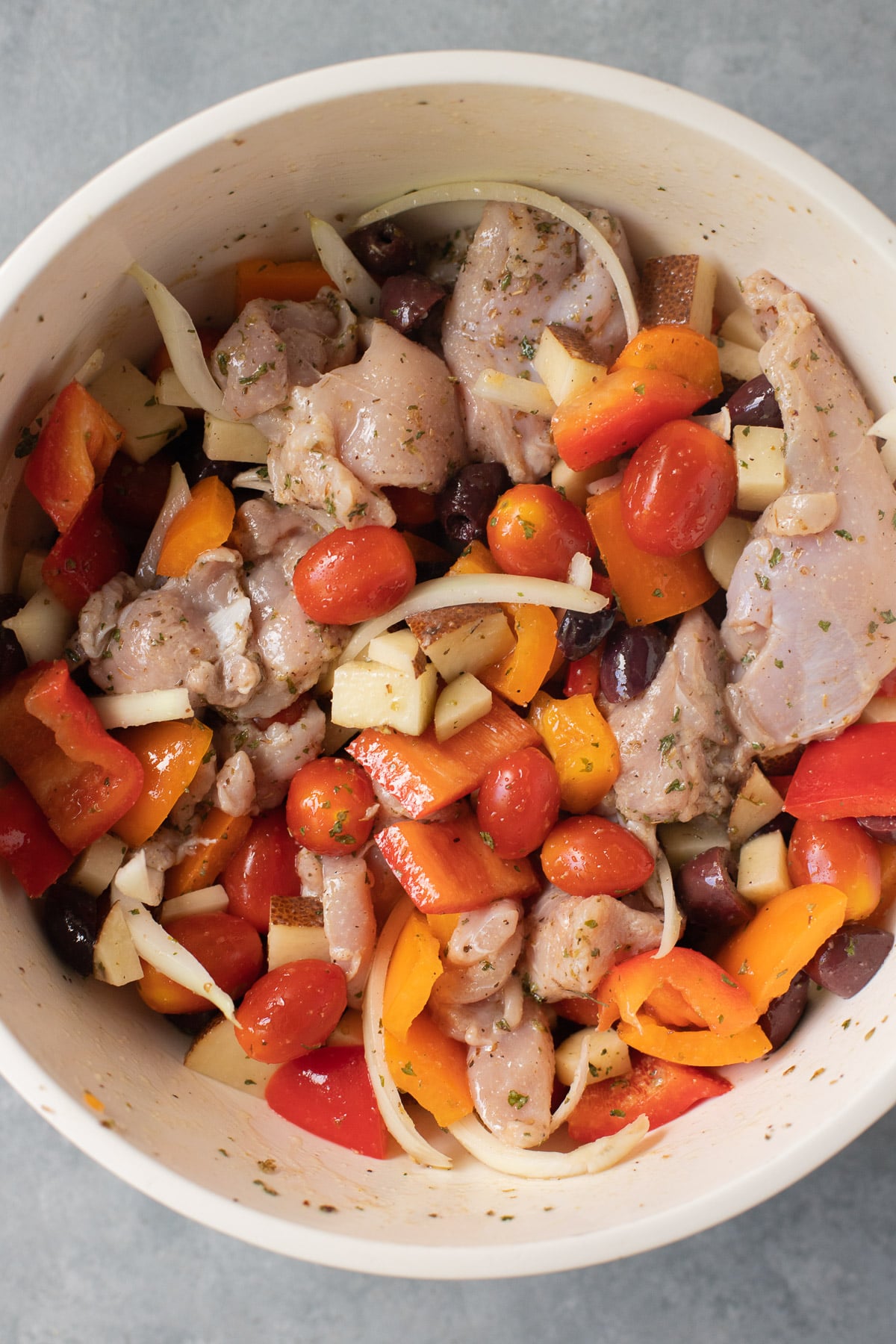 A large white bowl with raw chicken and veggies tossed in a marinade for greek chicken thighs