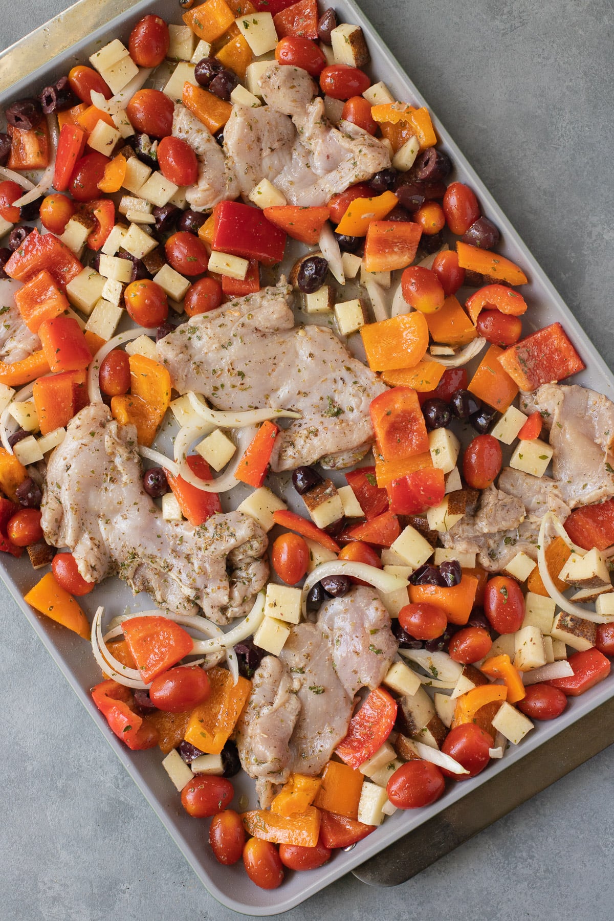 A sheet pan of uncooked greek seasoned chicken thighs and veggies