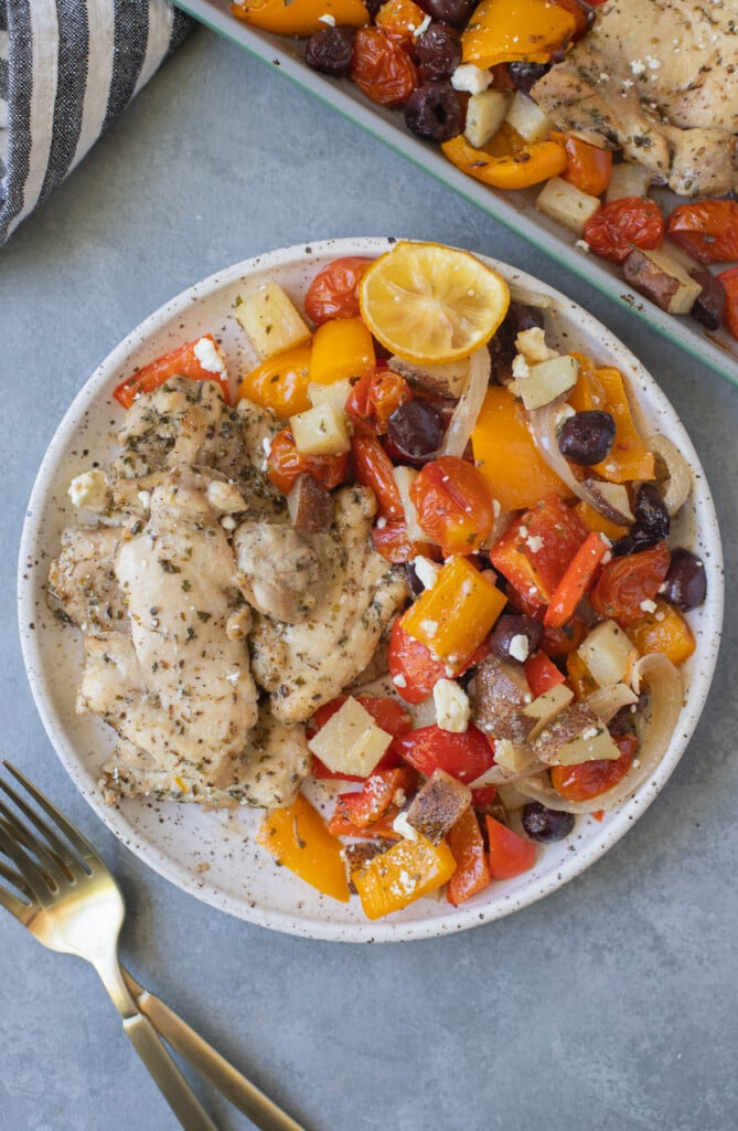 Greek Chicken Thighs and veggies on a white plate with gold forks and a sheet pan on the side