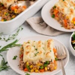 A white plate with healthy shepherd's pie with a fork on the plate and another plate and baking dish of the pie in the background