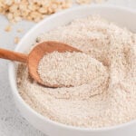 A white bowl of oat flour with a wooden scoop in it with rolled oats in the background