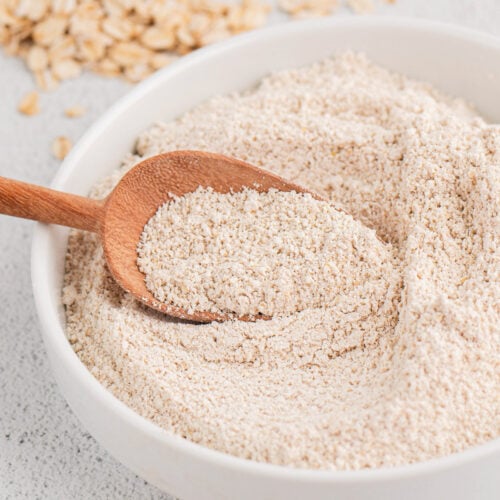 A white bowl of oat flour with a wooden scoop in it with rolled oats in the background