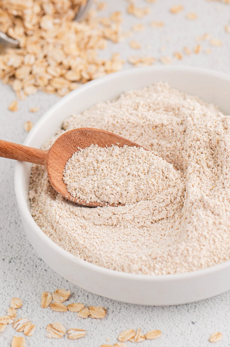 A white bowl of oat flour with a wooden scoop in it with rolled oats in the background
