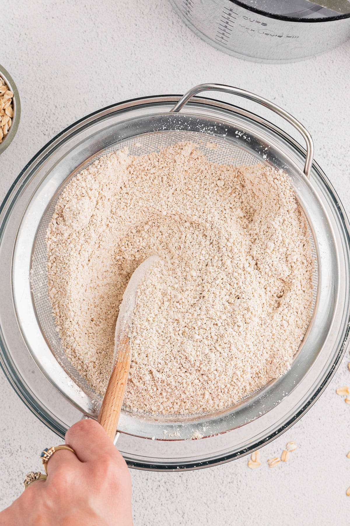 Hand holding a wooden spoon, pushing homemade oat flour through a fine mesh strainer into a glass mixing bowl.