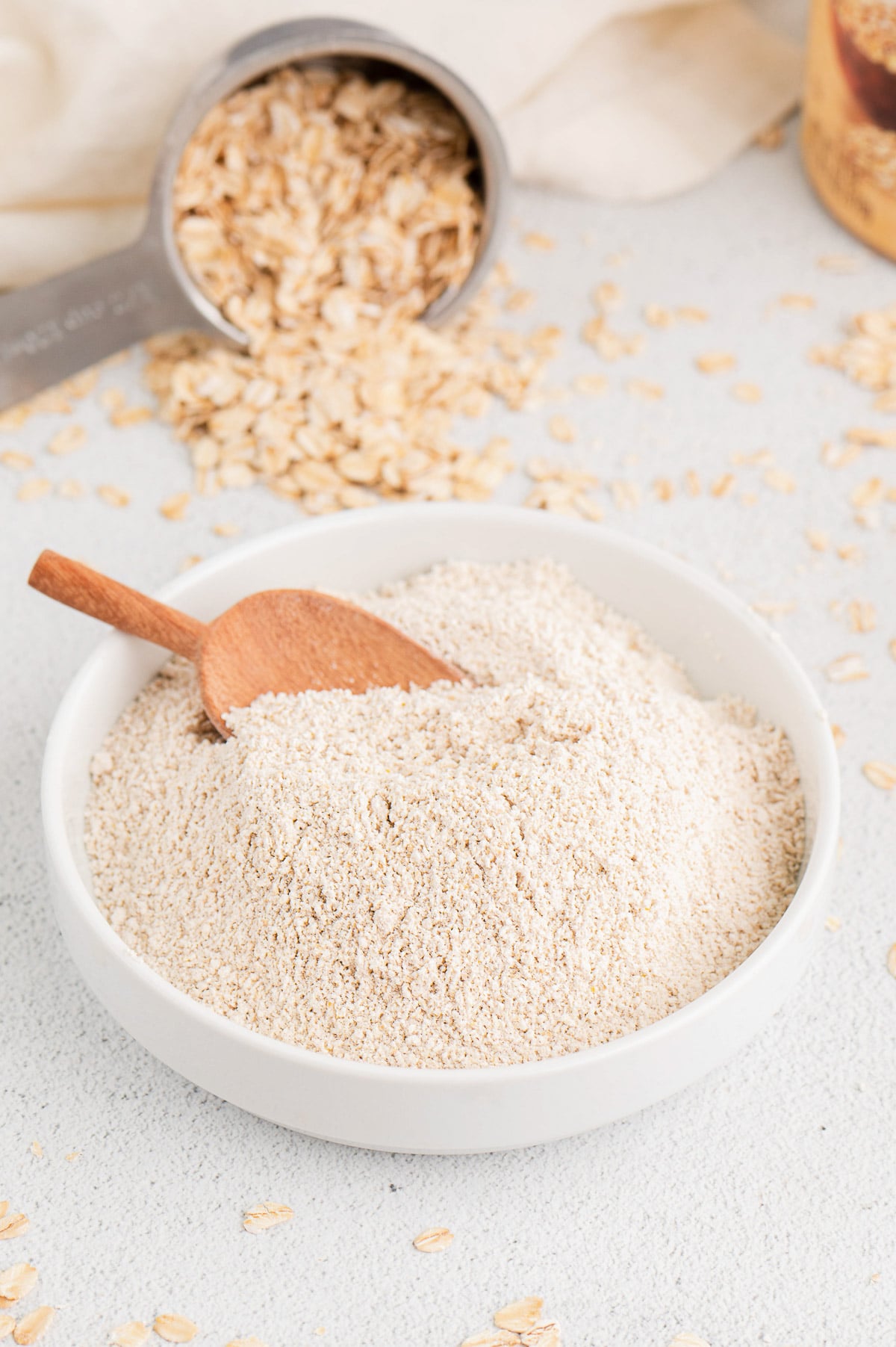 A white bowl of oat flour with a wooden scoop in it and a measuring up of old fashioned rolled oats in the background.
