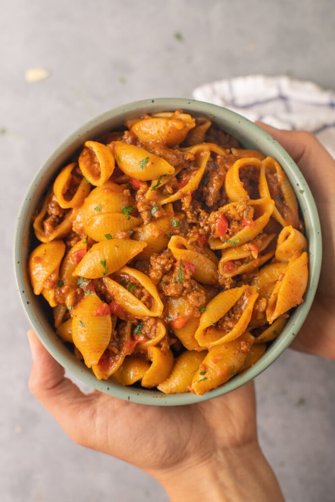 Two hands holding up a bowl of cheesy taco pasta with a grey background