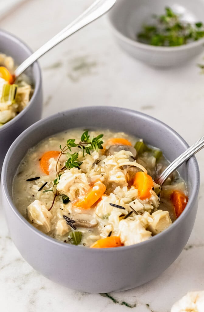 A grey bowl of turkey rice soup with a spoon in it with another bowl of soup and spices in the background on a marble top