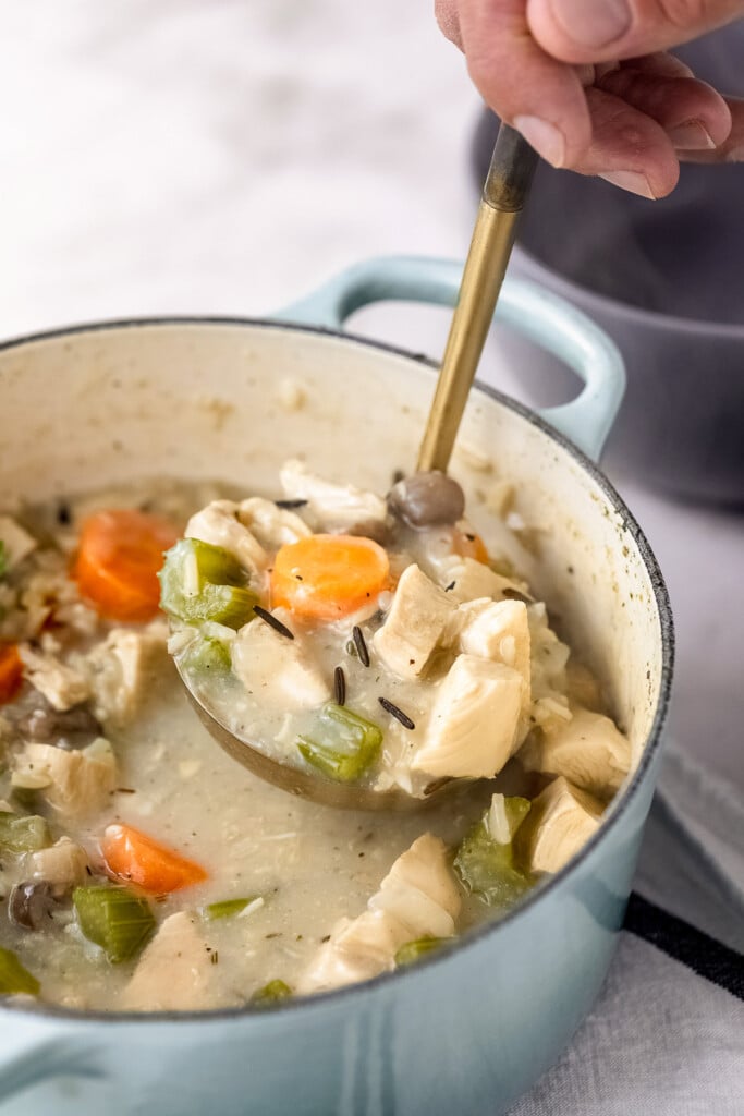 A large blue pot of turkey wild rice soup with a ladle taking a large scoop out of the soup on a marble background
